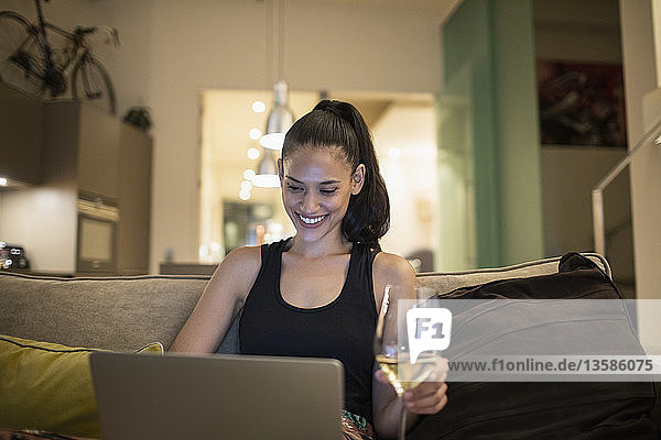 Lächelnde Frau  die einen Laptop benutzt und Weißwein auf einem Sofa in einer Wohnung trinkt