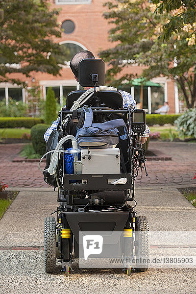 Rückansicht eines Mannes mit Duchenne-Muskeldystrophie in einem motorisierten Rollstuhl auf der Straße