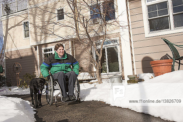 Frau mit Multipler Sklerose in einem Rollstuhl mit einem Diensthund vor ihrem Haus