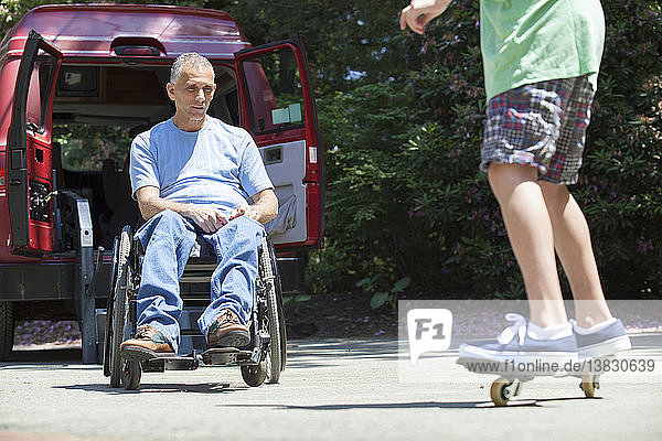Mann mit Querschnittslähmung im Rollstuhl beobachtet seinen Sohn auf dem Skateboard