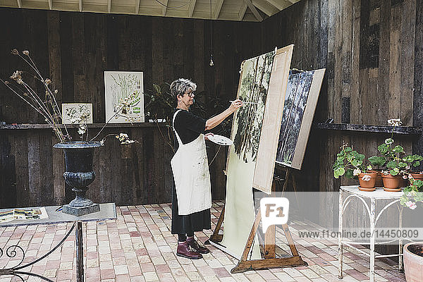 Ältere Frau mit Brille  schwarzem Oberteil und weißer Schürze steht im Atelier und arbeitet an der Bemalung von Bäumen im Wald.