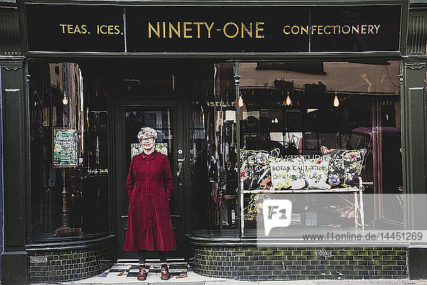 Lächelnde ältere Frau mit Brille und rotem Kleid steht vor einem Einrichtungshaus und schaut in die Kamera.