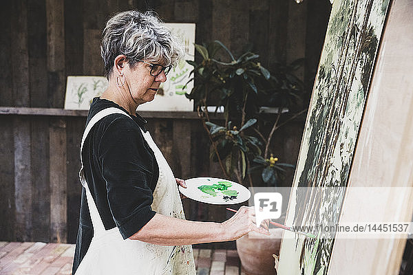 Ältere Frau mit Brille schwarzem Oberteil und weißer Schürze steht im Atelier und arbeitet an der Bemalung von Bäumen im Wald.