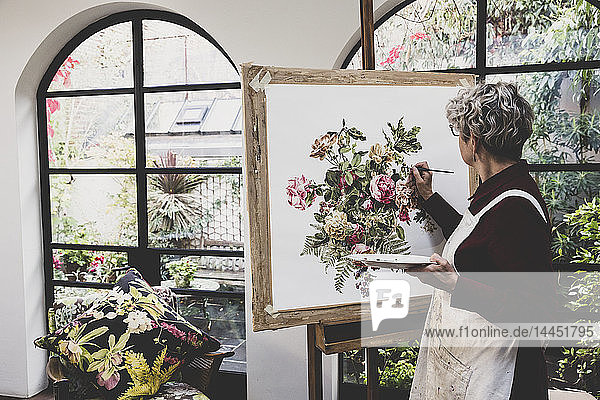 Ältere Frau mit Brille  rotem Kleid und weißer Schürze steht im Atelier und arbeitet an der Malerei von rosa Teerosen  Blättern  Beeren und anderen Blumen.