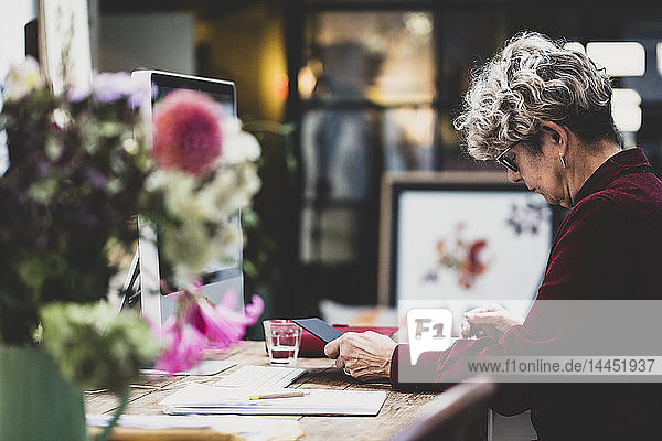 Ältere Frau mit Brille und rotem Kleid sitzt an einem Holztisch und schaut auf ein digitales Tablett.