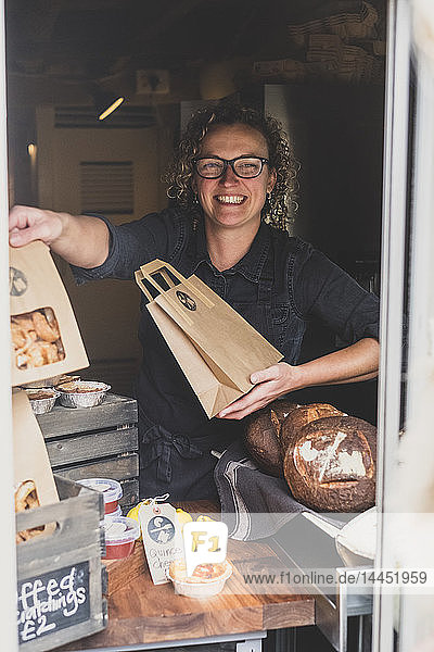 Lächelnde Frau mit Brille  die in einer Bäckerei steht und eine braune Einkaufstasche aus Papier in der Hand hält.