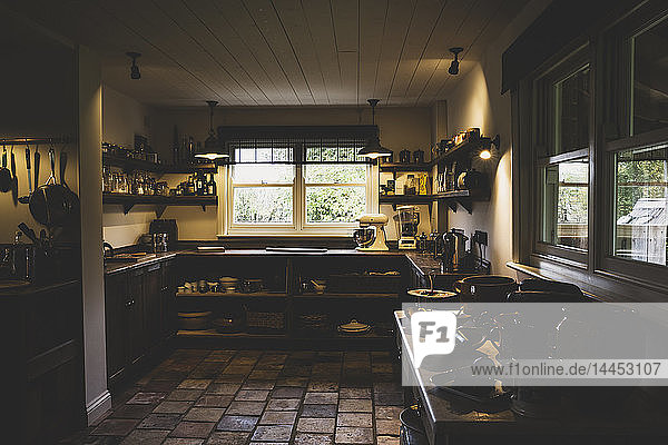 Innenansicht der Küche mit Steinfliesenboden  Holzdecke und zwei Schiebefenstern  antiken Holzschränken und Wandregalen.