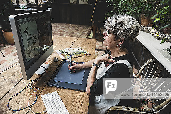 Ältere Frau mit Brille  schwarzem Oberteil und weißer Schürze sitzt an einem Holztisch und arbeitet am Desktop-Computer.