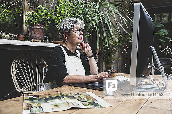 Ältere Frau mit Brille  schwarzem Oberteil und weißer Schürze sitzt an einem Holztisch und arbeitet am Desktop-Computer.