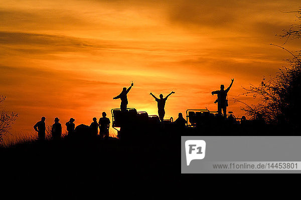 Eine Silhouette von Menschen und einem Fahrzeug  Menschen mit Händen in der Luft  vor Sonnenuntergang  orangefarbenem und gelbem Himmel