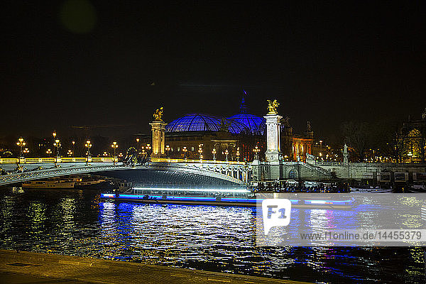 Nachtansicht eines Flugbootes  das die Seine vor dem Grand Palais mit seiner beleuchteten Kuppel und der beleuchteten Brücke Alexandre III in Paris  Frankreich  überquert.