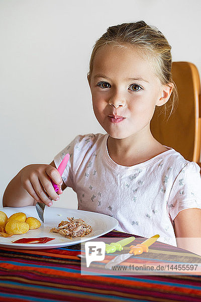 Ein hübsches  lächelndes kleines Mädchen  das am Tisch sitzend Kartoffeln und Hühnchen isst.