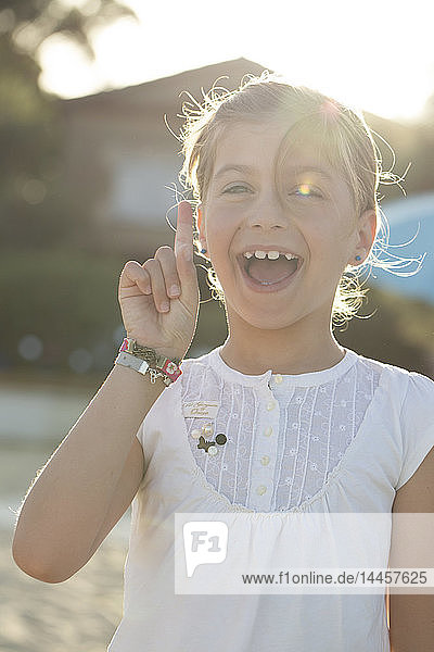 Hübsches ausdrucksstarkes Mädchen sehr glücklich mit einer Hand neben dem Gesicht  den Zeigefinger draußen im Sommer bei Sonnenuntergang erhoben.