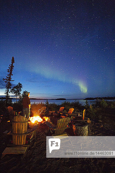 A man views the Northern Lights (aurora borealis) by a camp fire by Lake Egenolf in northern Manitoba  Canada  North America