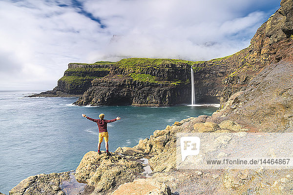Mann auf Klippen mit offenen Armen  der den Gasadalur-Wasserfall bewundert  Insel Vagar  Färöer Inseln  Dänemark