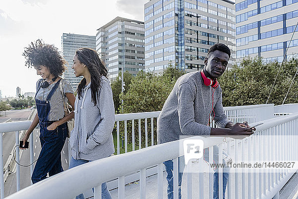 Junger Mann mit Kopfhörern steht auf der Brücke  benutzt ein Smartphone  junge Leute gehen vorbei