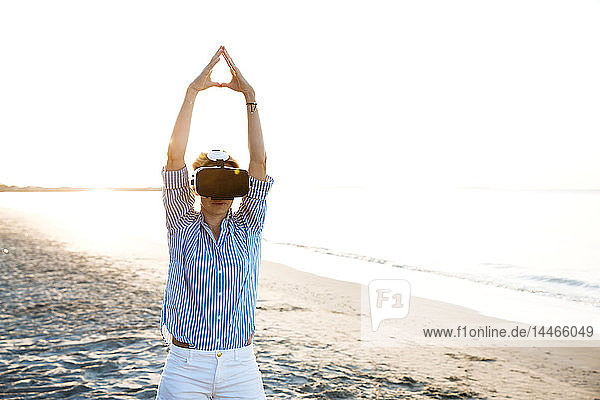 Blonde Frau macht eine Art Yoga-Übungen an einem Strand in Thailand mit einer 3D-Brille für virtuelle Realität