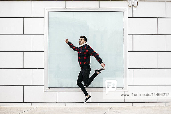 Junger Mann in legerer Kleidung springt mit einer weißen Wand und einem großen Fenster im Hintergrund