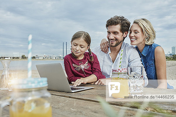 Deutschland  Düsseldorf  glückliche Familie mit Tochter mit Laptop auf Holztisch am Rheinufer