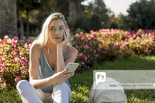 Lächelnde junge Frau sitzt im Park mit Handy und Ohrstöpseln