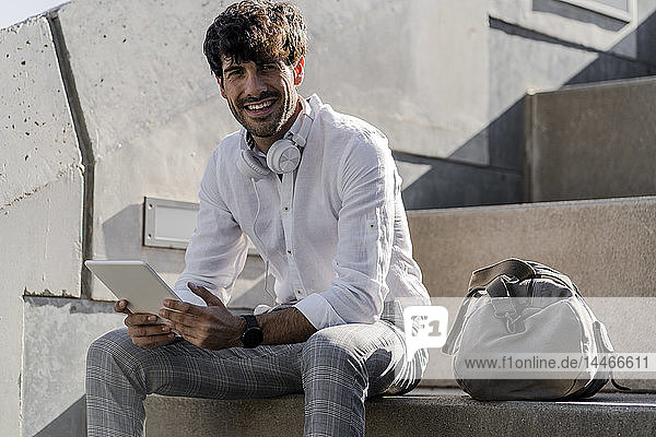 Portrait of smiling young man sitting on stairs outdoors using tablet