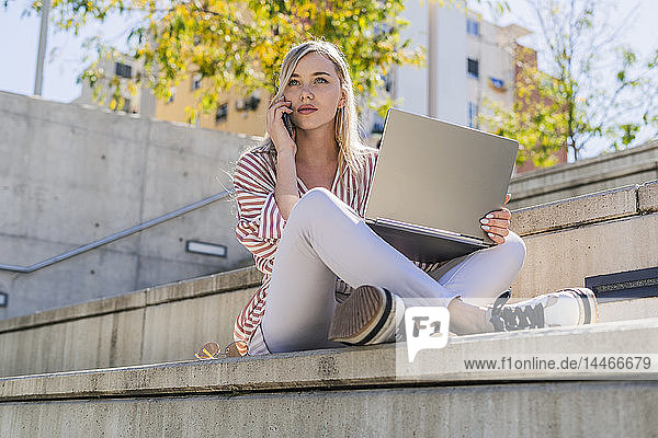 Porträt einer blonden jungen Frau am Telefon  die mit einem Laptop auf einer Treppe im Freien sitzt