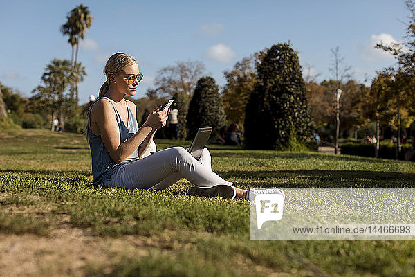 Junge Frau sitzt im Park und benutzt Handy und Laptop