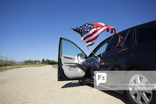 Mädchen mit amerikanischer Flagge sitzt im Auto auf einem Weg in abgelegener Landschaft