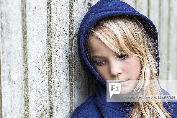 Portrait of sad blond girl wearing blue hooded jacket leaning against wooden wall