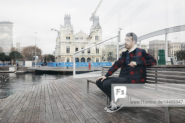 Young man wearing casual clothes sitting on a bench at harbour