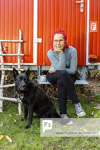 Porträt einer zufriedenen älteren Frau mit rot gefärbten Haaren  die mit ihrem Hund vor einem roten Wohnwagen im Garten sitzt