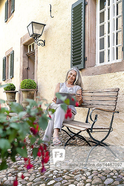 Frau mit langen grauen Haaren sitzt mit Tablette auf Bank im Landhaus