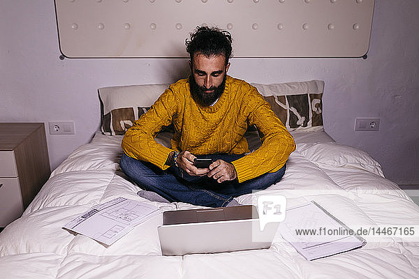 Junger Mann arbeitet zu Hause im Bett mit Handy  Laptop und Dokumenten