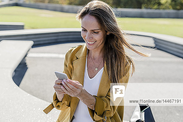 Smiling woman with bag sitting on a bench in the city using cell phone