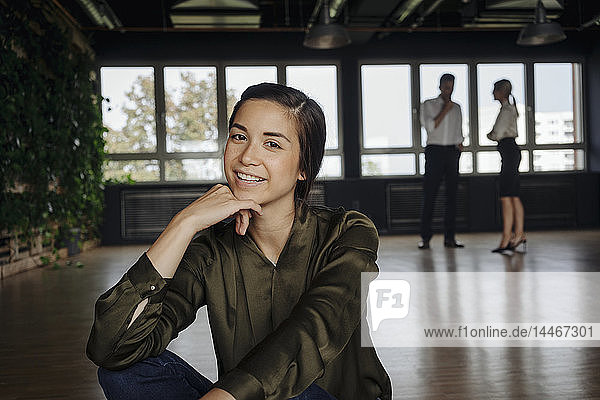 Lächelnde Frau sitzt im Büro auf dem Boden mit zwei Personen im Hintergrund