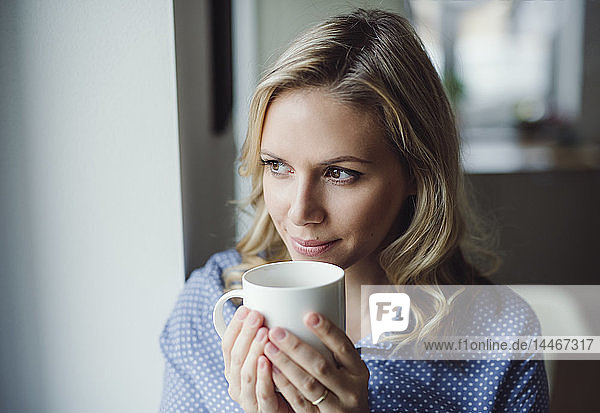Lächelnde Frau hält zu Hause eine Tasse Kaffee am Fenster