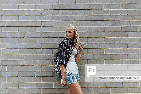 Lächelnde junge Frau mit Handy an Ziegelmauer