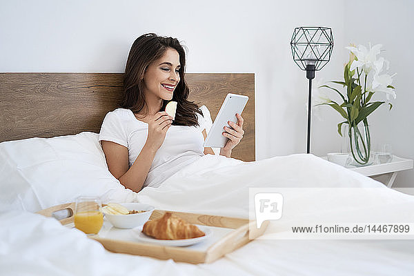 Junge Frau frühstückt im Bett  benutzt digitales Tablett  liest