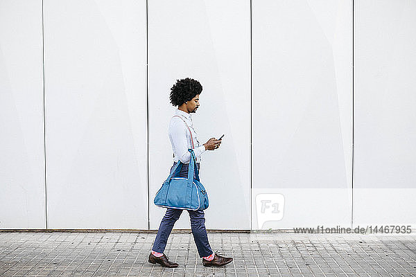 Mann mit Tasche geht vor einer weißen Wand  während er auf sein Handy schaut