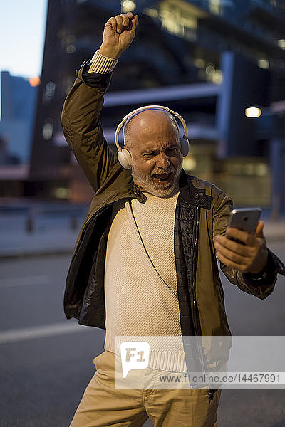 Spain  Barcelona  excited senior man with headphones and cell phone in the city at dusk
