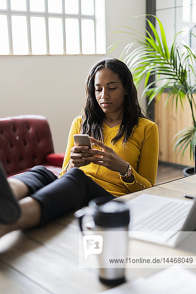 Junge Geschäftsfrau sitzt mit den Füßen auf dem Schreibtisch und benutzt ein Mobiltelefon