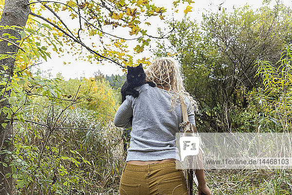 Rückenansicht einer Bogenschützin mit Bogen  die in der Natur eine schwarze Katze auf der Schulter trägt