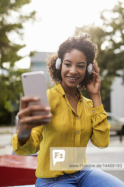Porträt einer lächelnden Frau  die mit Kopfhörern Musik hört und sich mit einem Smartphone selbstständig macht