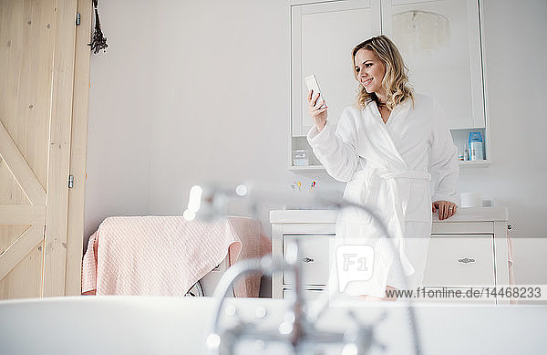 Lächelnde Frau im Bademantel im Badezimmer zu Hause mit Handy in der Hand