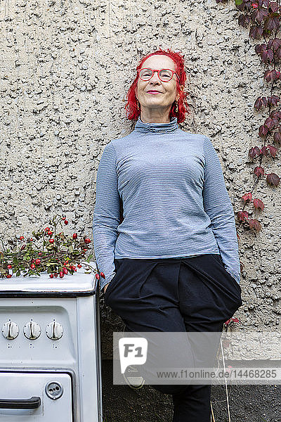 Porträt einer lächelnden älteren Frau mit rot gefärbtem Haar  die sich an die Hausfront lehnt