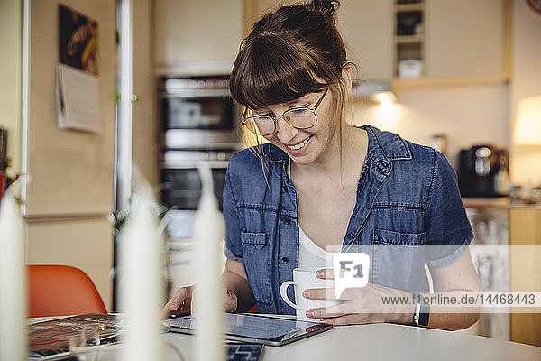 Lächelnde Frau sitzt mit einer Tasse Kaffee am Tisch in der Küche und benutzt Tabletten