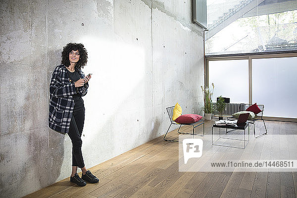 Smiling woman with cell phone leaning against concrete wall in a loft