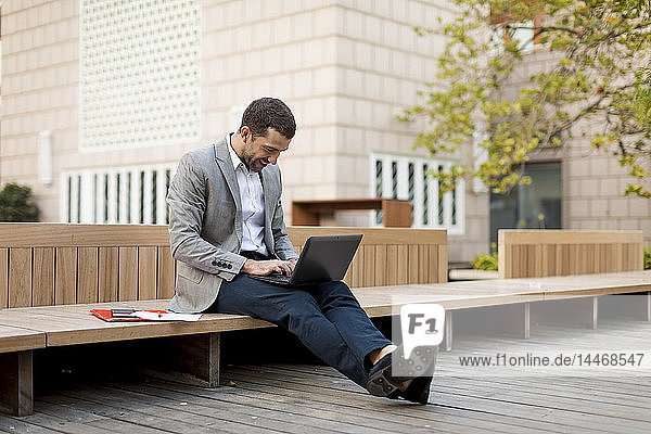 Lächelnder Geschäftsmann sitzt mit Laptop auf einer Bank