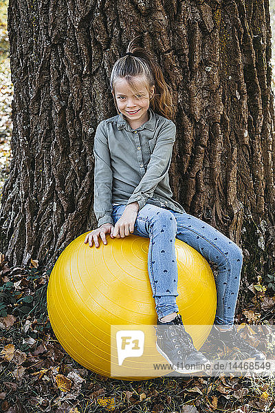 Porträt eines lächelnden Mädchens  das auf einem gelben Gymnastikball sitzt und an einen Baumstamm gelehnt ist
