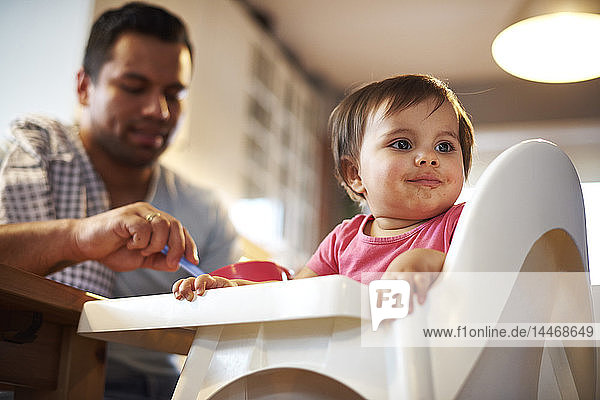 Portrait of baby girl sitting in high chair at home with father in background